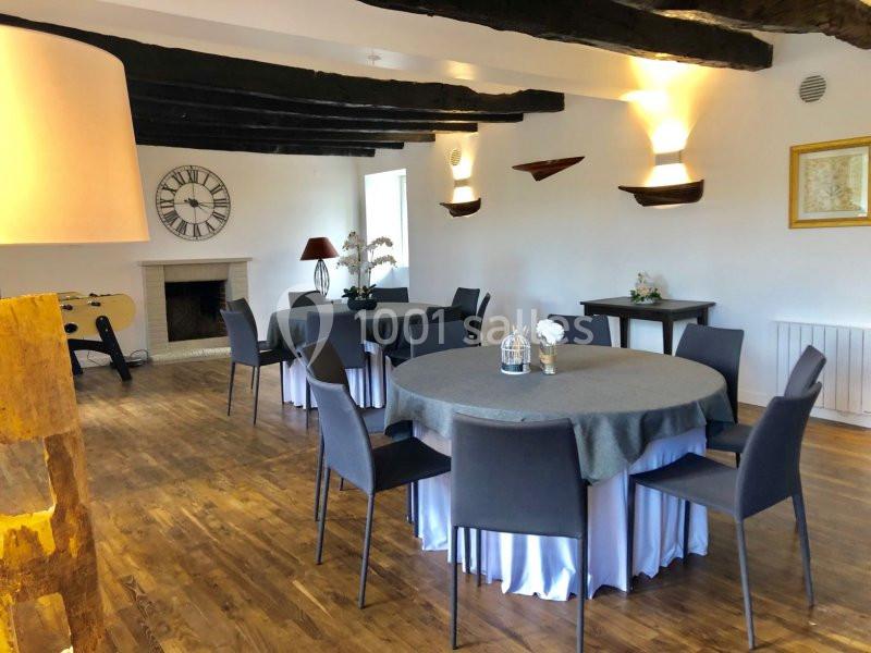 Salle lumineuse avec parquet en bois, tables rondes dressées, chaises noires, cheminée et décoration murale.