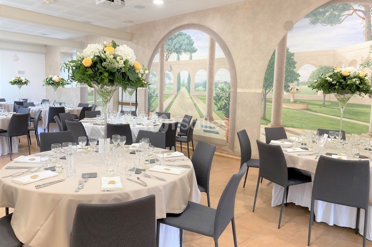 Salle de réception élégante avec tables rondes dressées, décorée de fresques murales représentant un jardin méditerranéen.