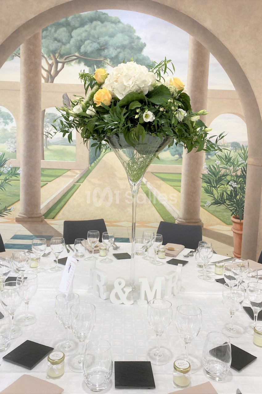 Centre de table floral avec roses jaunes et blanches dans un vase haut, entouré de vaisselle sur une table dressée.