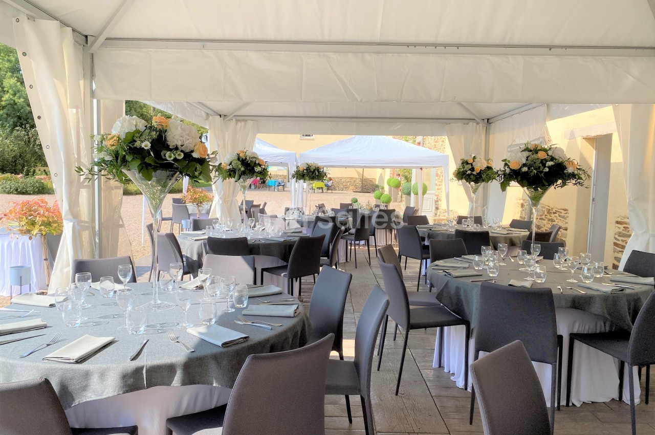 Salle de réception en extérieur sous une tente, avec tables dressées, nappes grises et décorations florales.