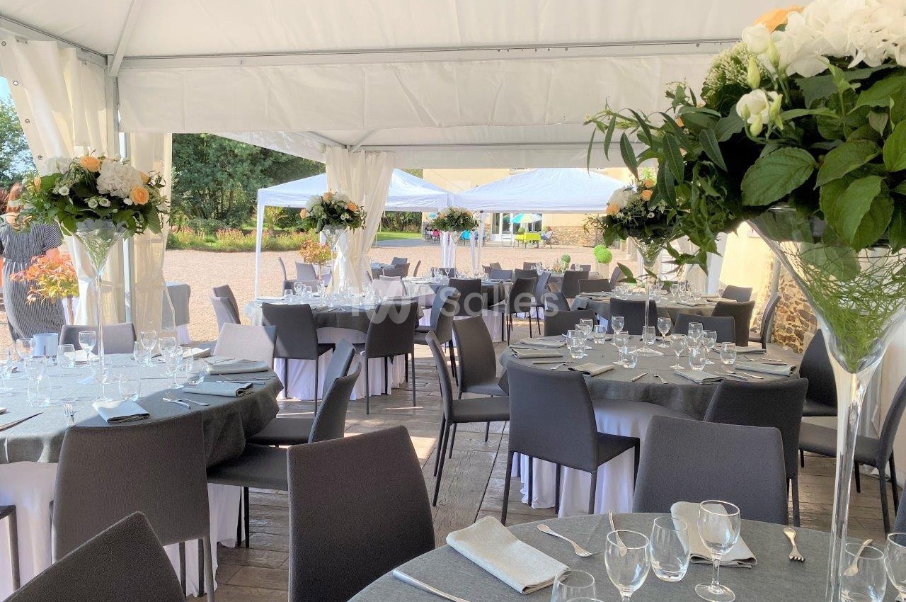 Salle de réception sous tente avec tables rondes dressées, nappes grises, chaises noires et décorations florales.