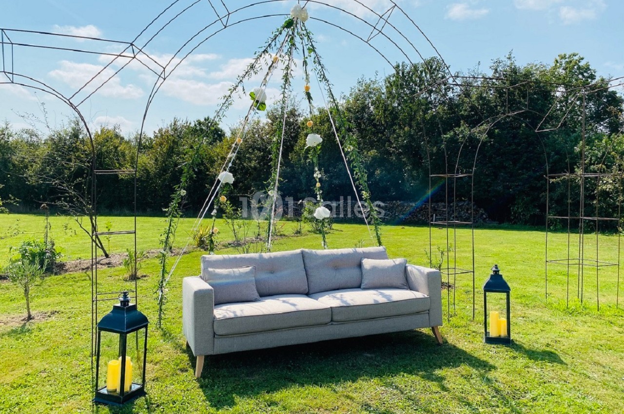 Canapé gris disposé sous une arche métallique décorée de fleurs et feuillages, dans un jardin ensoleillé.