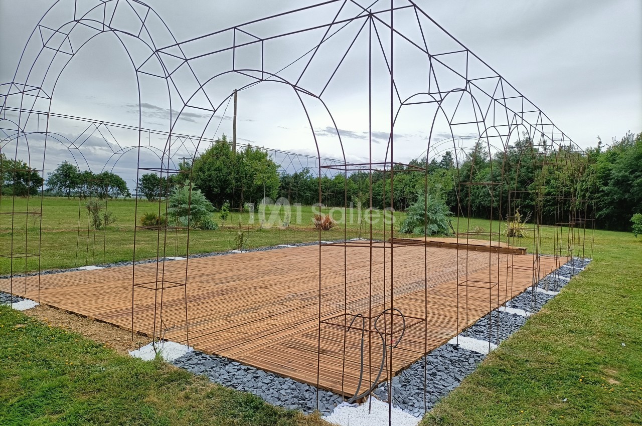 Structure métallique en arc installée sur une plateforme en bois, entourée de pelouse et d'arbres sous un ciel nuageux.