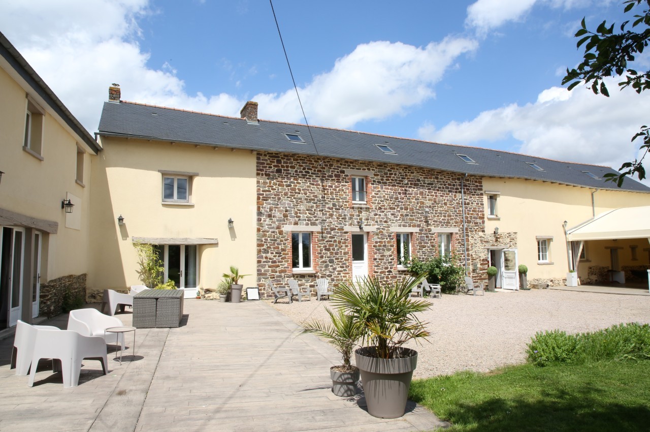 Façade d'une maison en pierre et crépi avec une cour aménagée, mobilier d'extérieur et ciel partiellement nuageux.