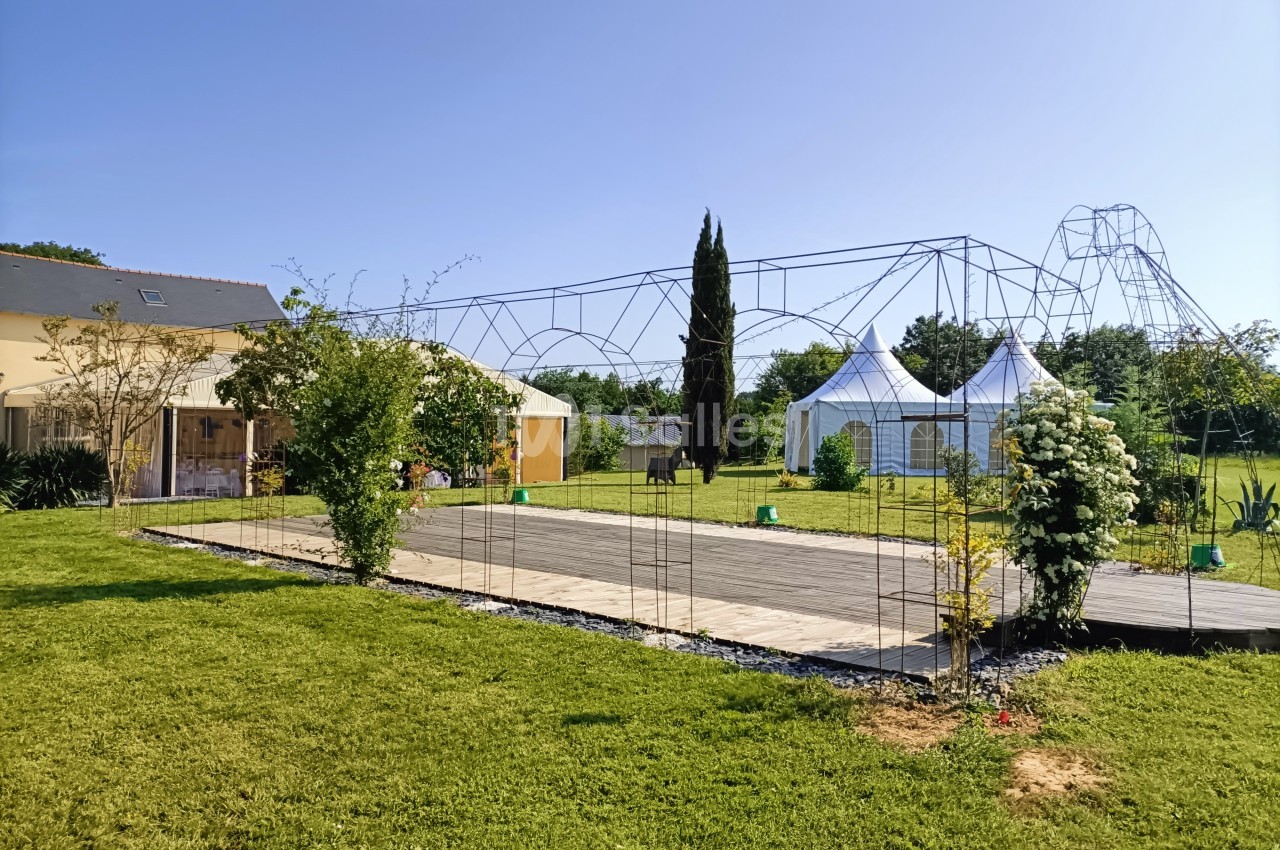 Jardin avec pelouse, pergola métallique, maison à gauche et tentes blanches en arrière-plan par temps ensoleillé.