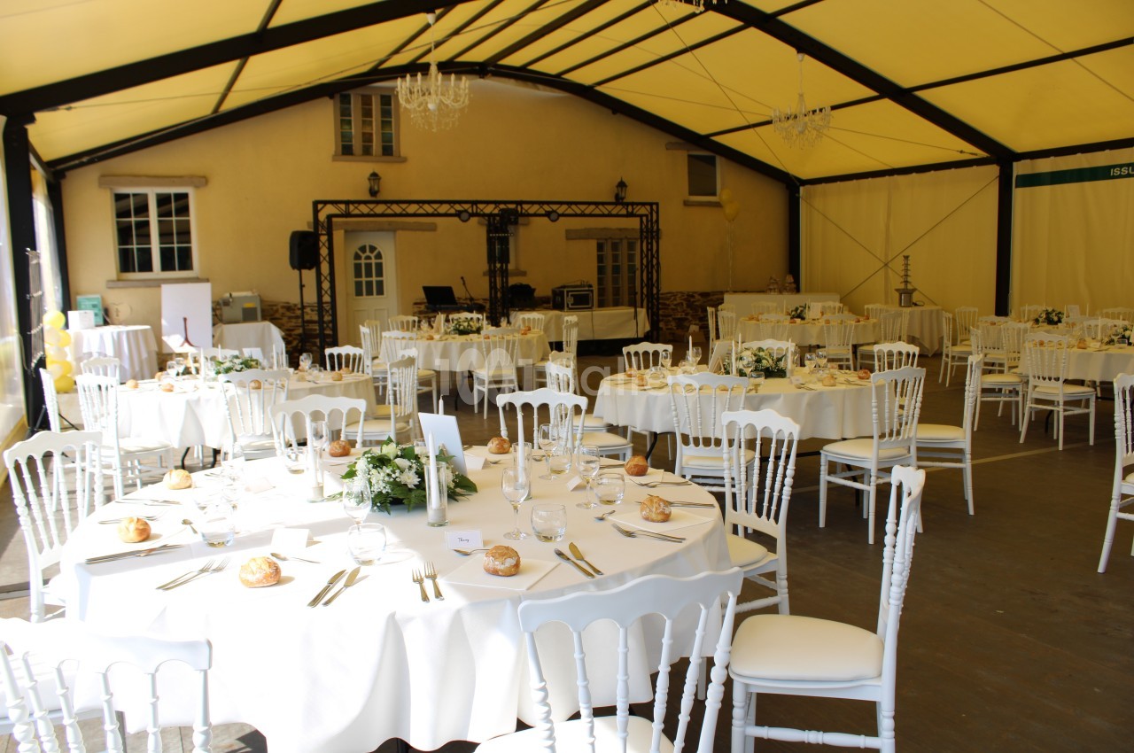Salle de réception sous une tente avec tables rondes dressées, nappes blanches, chaises assorties et décorations florales.