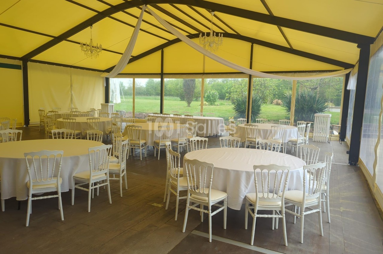 Salle de réception lumineuse avec tables rondes dressées, chaises blanches et vue sur un jardin verdoyant à travers des…