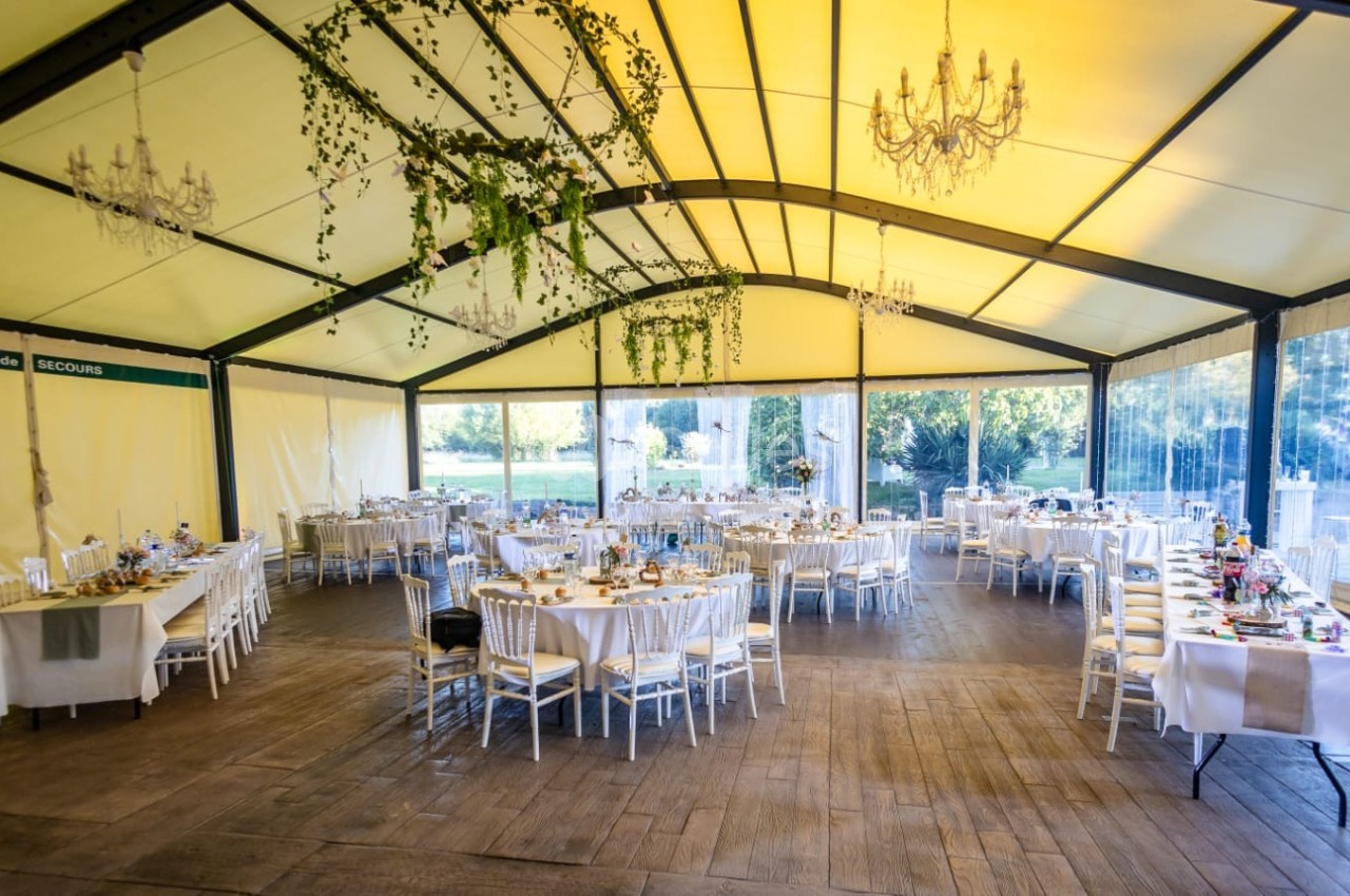 Salle de réception sous une tente avec tables décorées, chaises blanches et guirlandes végétales suspendues.
