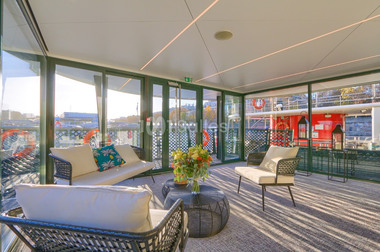 Salon lumineux avec mobilier en rotin, table basse et grandes baies vitrées donnant sur un bateau et un paysage urbain.