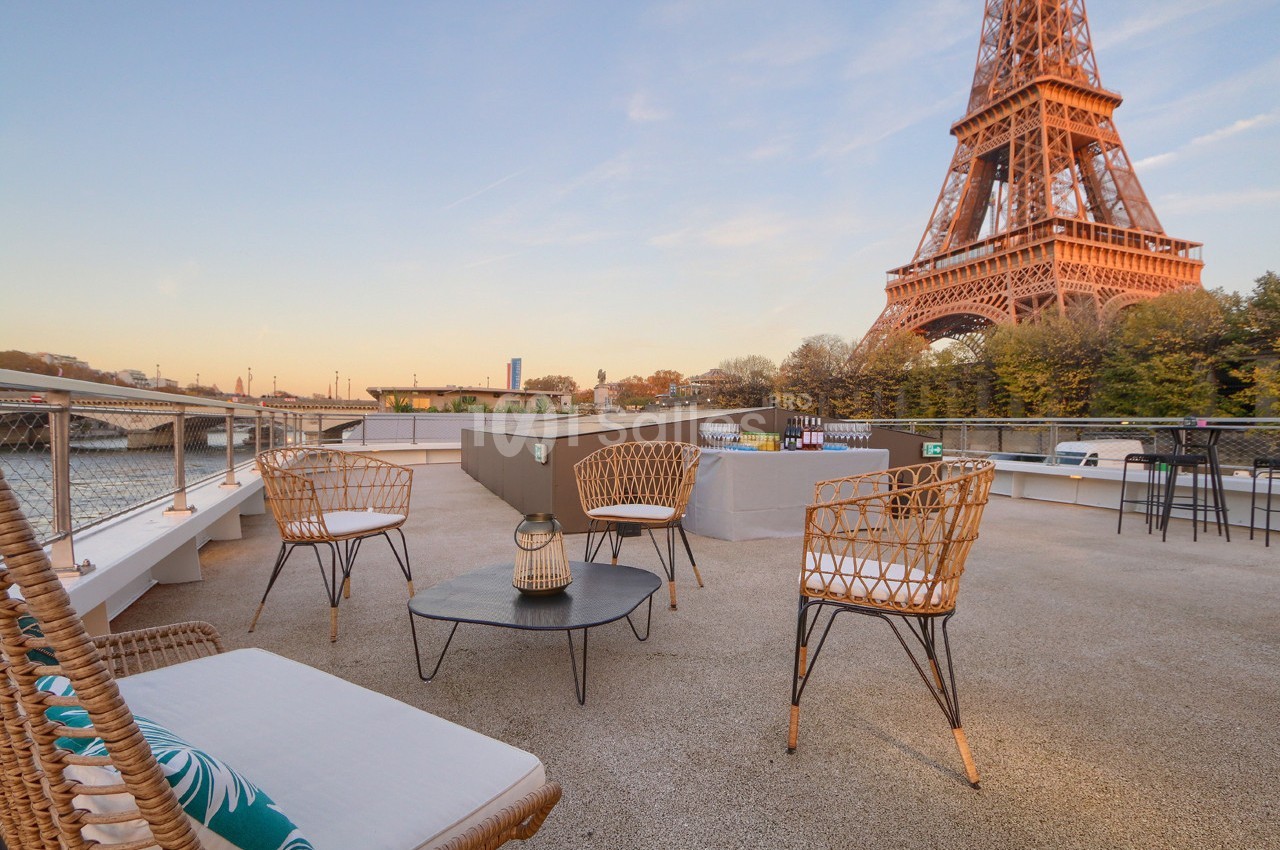 Terrasse avec mobilier en rotin, vue sur la Tour Eiffel et la Seine sous un ciel dégagé en fin de journée.