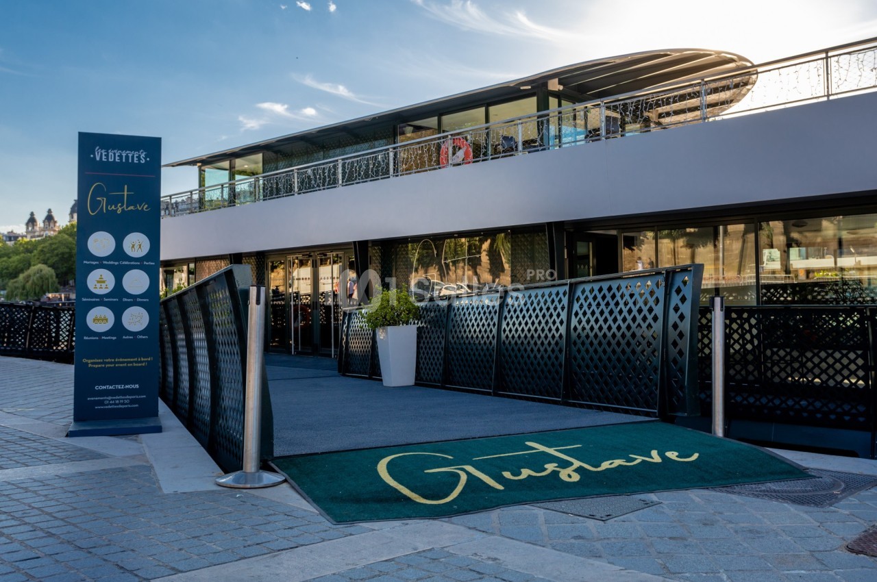 Entrée d'un restaurant flottant moderne avec une enseigne ’Gustave’, un tapis vert et un panneau d'information.