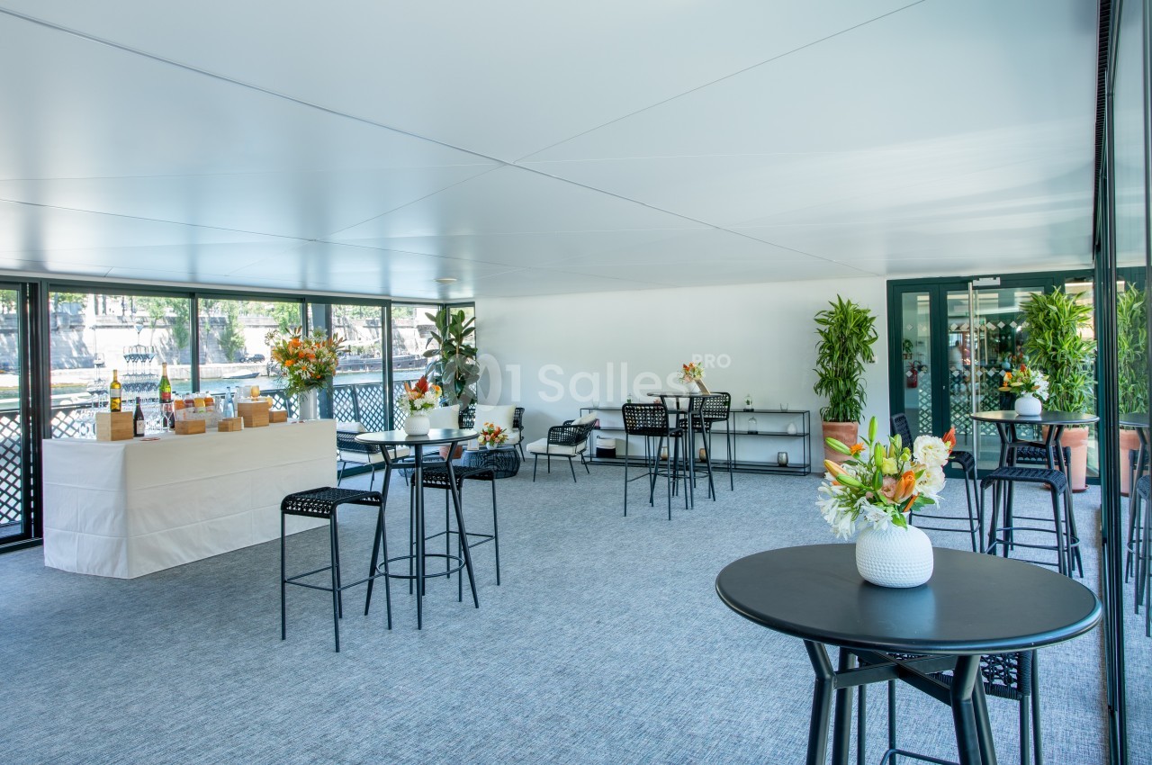 Salle lumineuse avec tables hautes, chaises noires, plantes décoratives et buffet dressé près d'une baie vitrée.