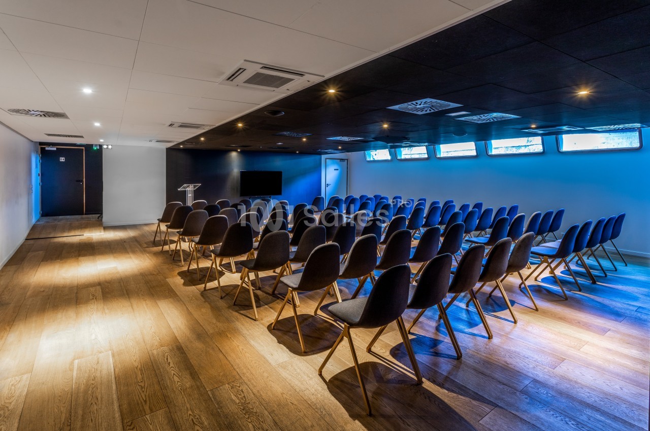 Salle de conférence vide avec rangées de chaises noires, parquet en bois et éclairage tamisé.