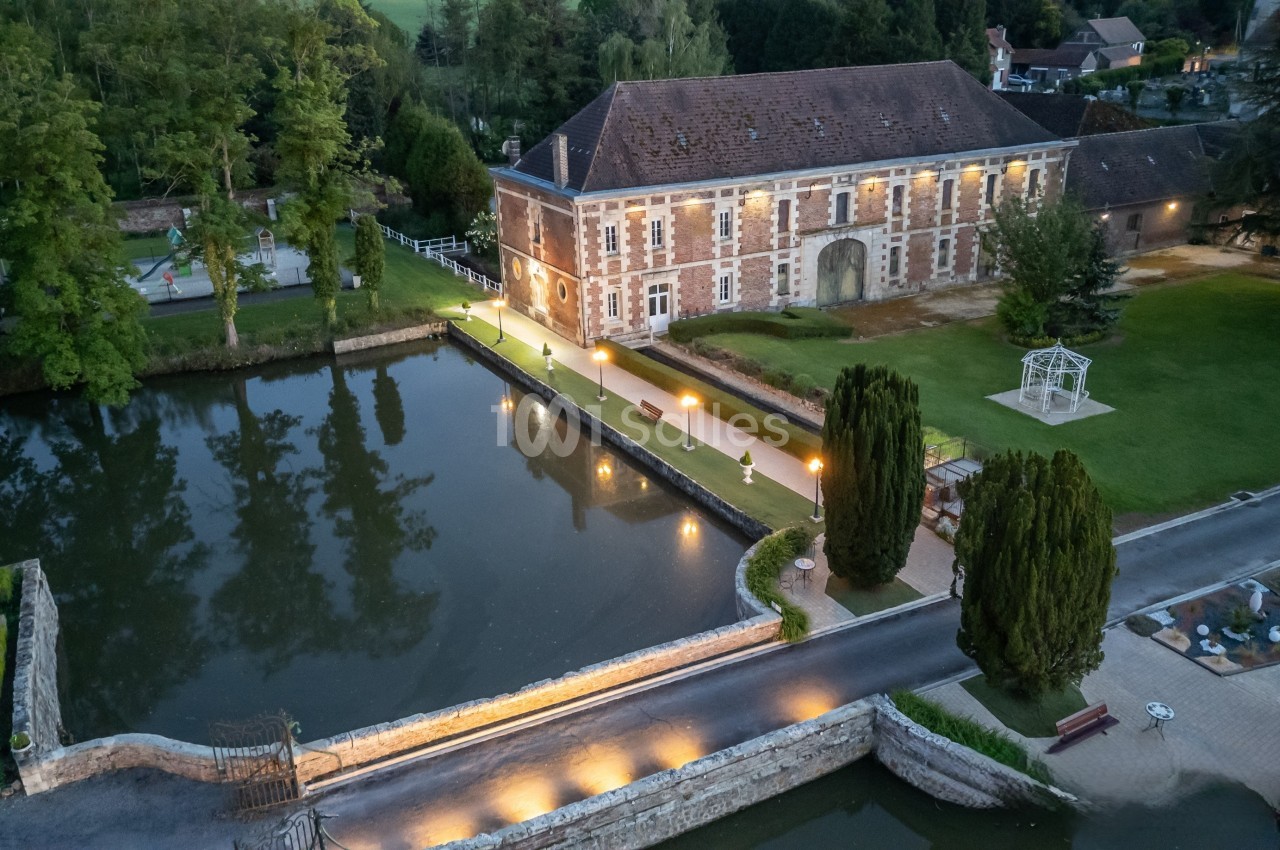 Vue aérienne d'un bâtiment ancien en briques près d'un plan d'eau, entouré de verdure et éclairé par des lampadaires.