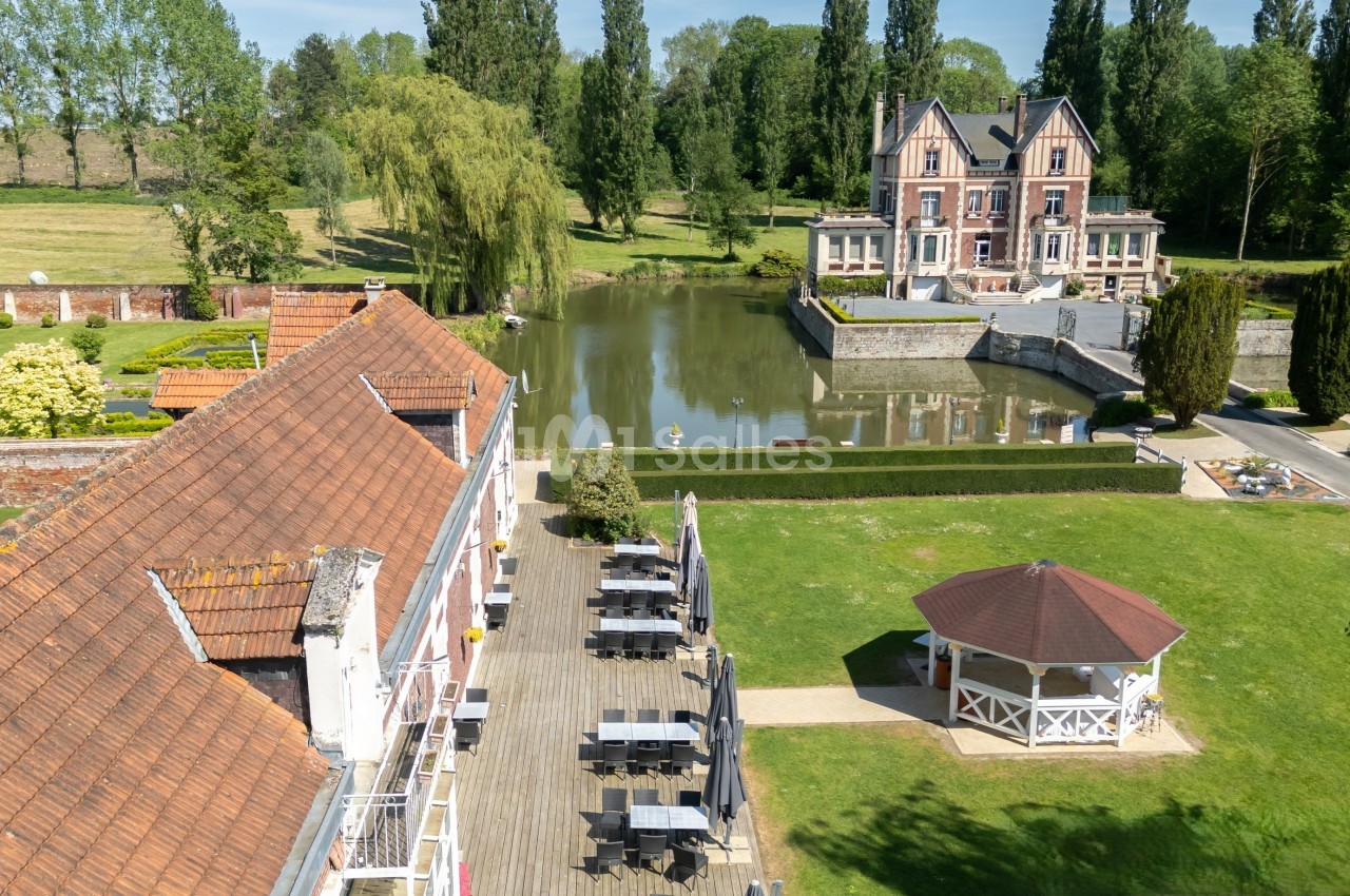 Vue aérienne d'un domaine avec un bâtiment en briques, un pavillon, un étang et un manoir entouré de verdure.