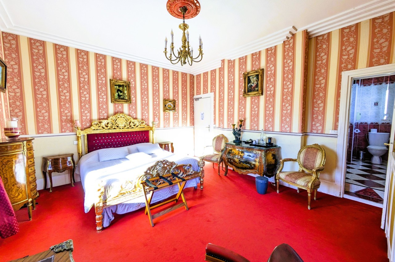 Chambre avec lit double, mobilier ancien, murs à rayures rouges et blanches, sol en moquette rouge, vue sur une salle de…