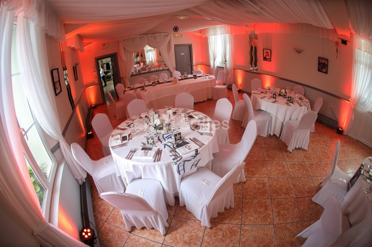 Salle de réception décorée avec des nappes blanches, des chaises couvertes et un éclairage tamisé rouge.