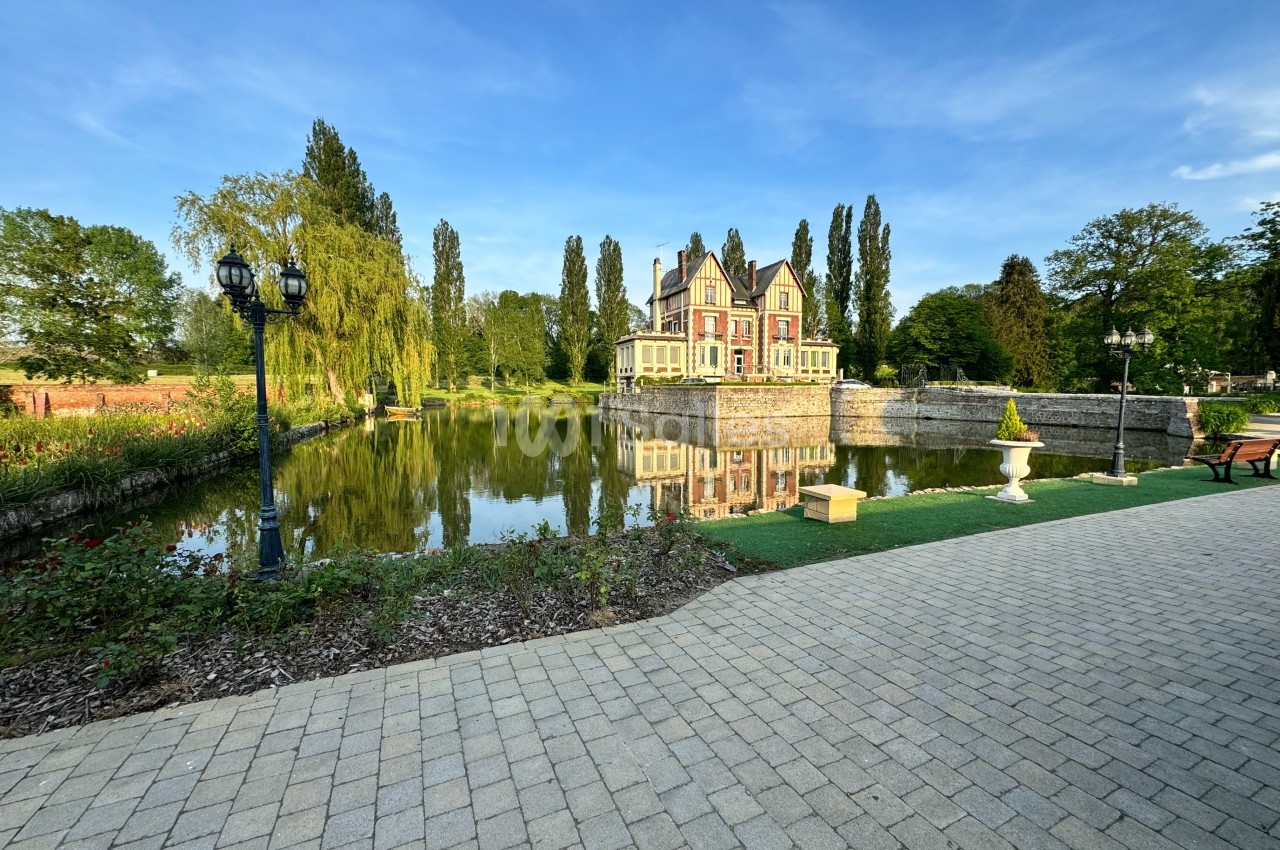 Manoir entouré d'arbres, reflété dans un étang, avec une allée pavée et un jardin au premier plan.