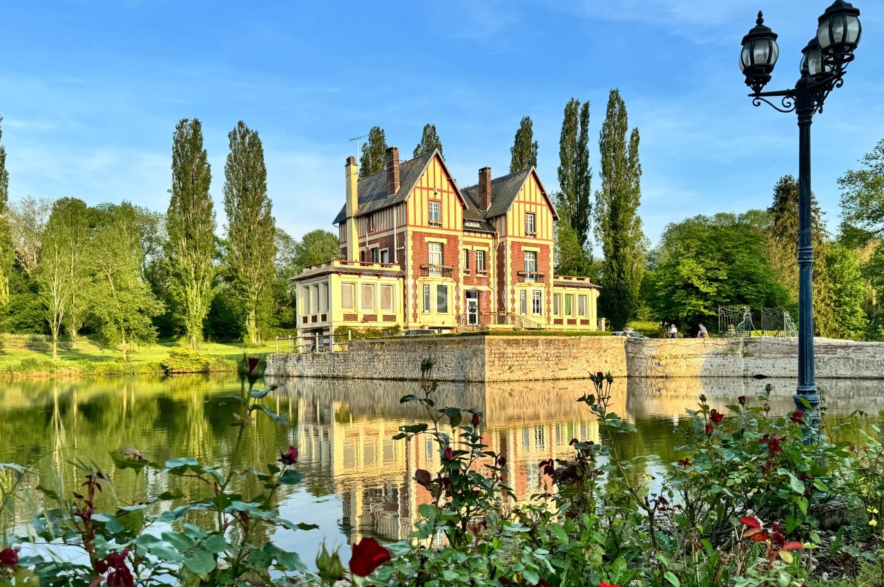 Manoir entouré d'arbres, reflété dans un étang calme, avec des roses au premier plan et un lampadaire à droite.