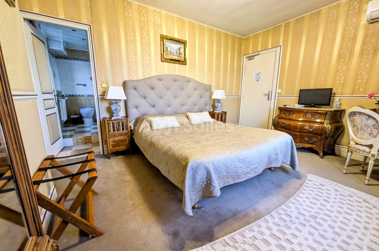 Chambre avec lit double, mobilier classique, tapis, et accès à une salle de bain visible en arrière-plan.