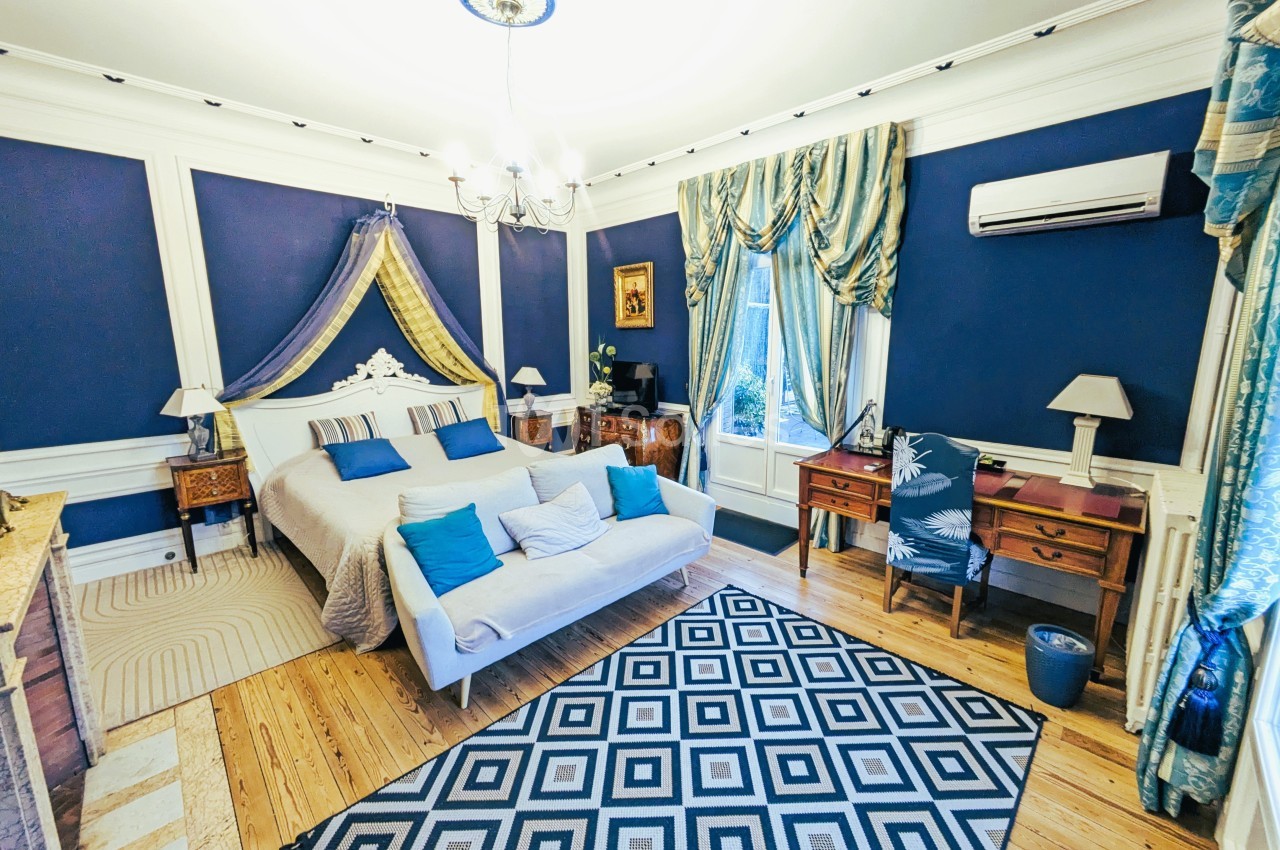 Chambre élégante avec murs bleus, lit double, mobilier classique, tapis géométrique et grande fenêtre avec rideaux.