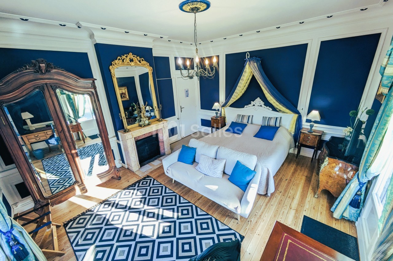 Chambre élégante avec murs bleus, lit double, cheminée, miroir doré, armoire en bois et tapis géométrique.