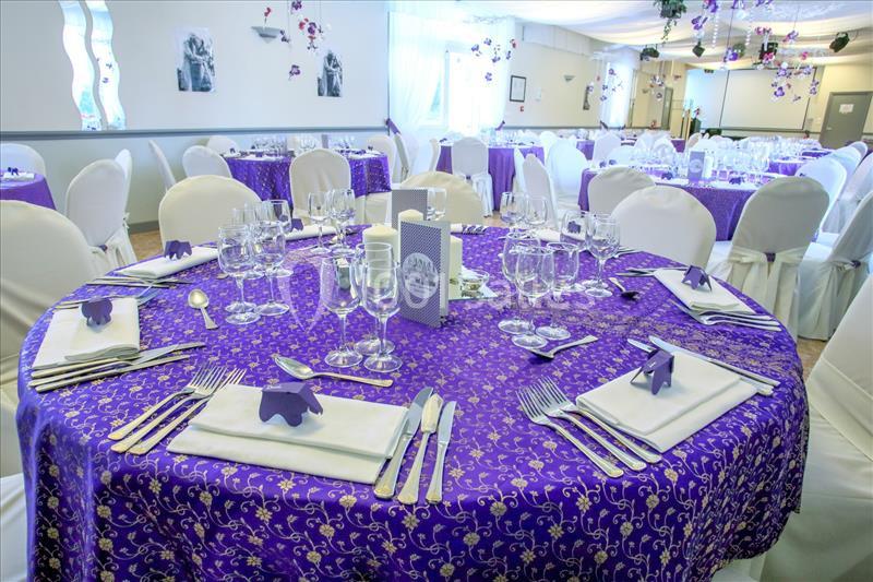 Salle de réception avec tables rondes dressées, nappes violettes ornées de motifs dorés et décorations élégantes.
