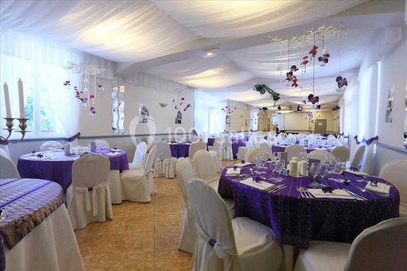 Salle de réception décorée avec des nappes violettes, des chaises blanches et des suspensions florales au plafond.