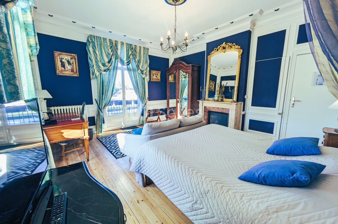 Chambre élégante avec murs bleus, lit double, cheminée, miroir doré, bureau en bois et grandes fenêtres avec rideaux.