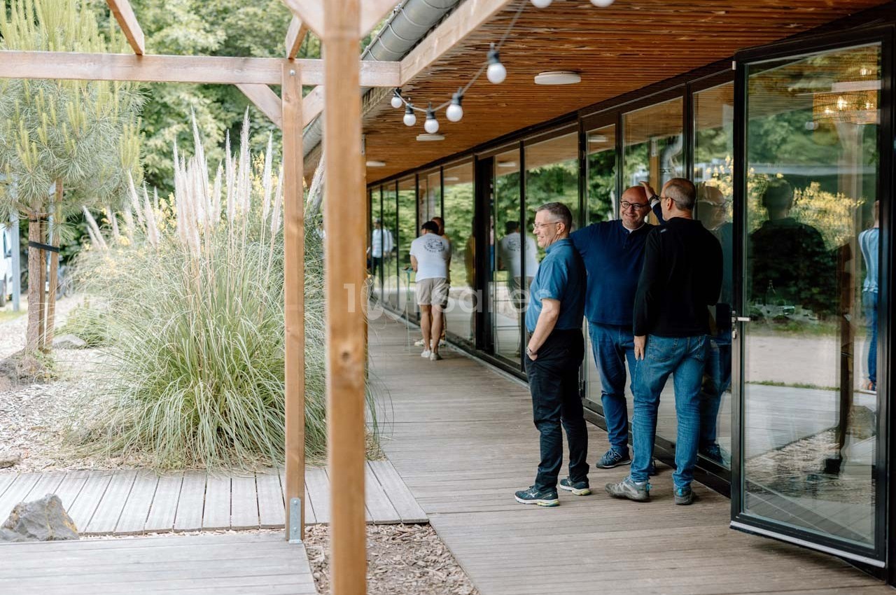Des personnes discutent sur une terrasse en bois bordée de baies vitrées, avec un jardin en arrière-plan.