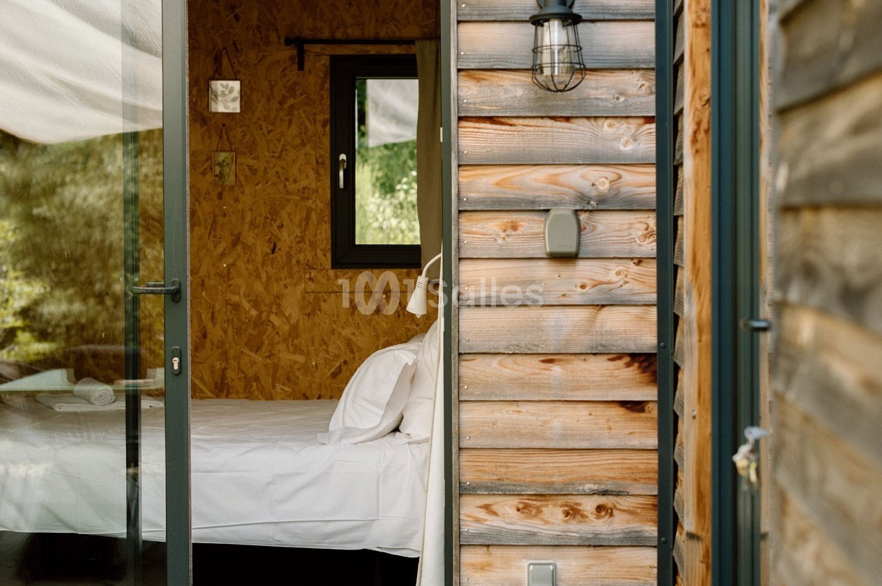 Vue partielle d'une chambre avec lit blanc, mur en bois et fenêtre, dans un cadre naturel.