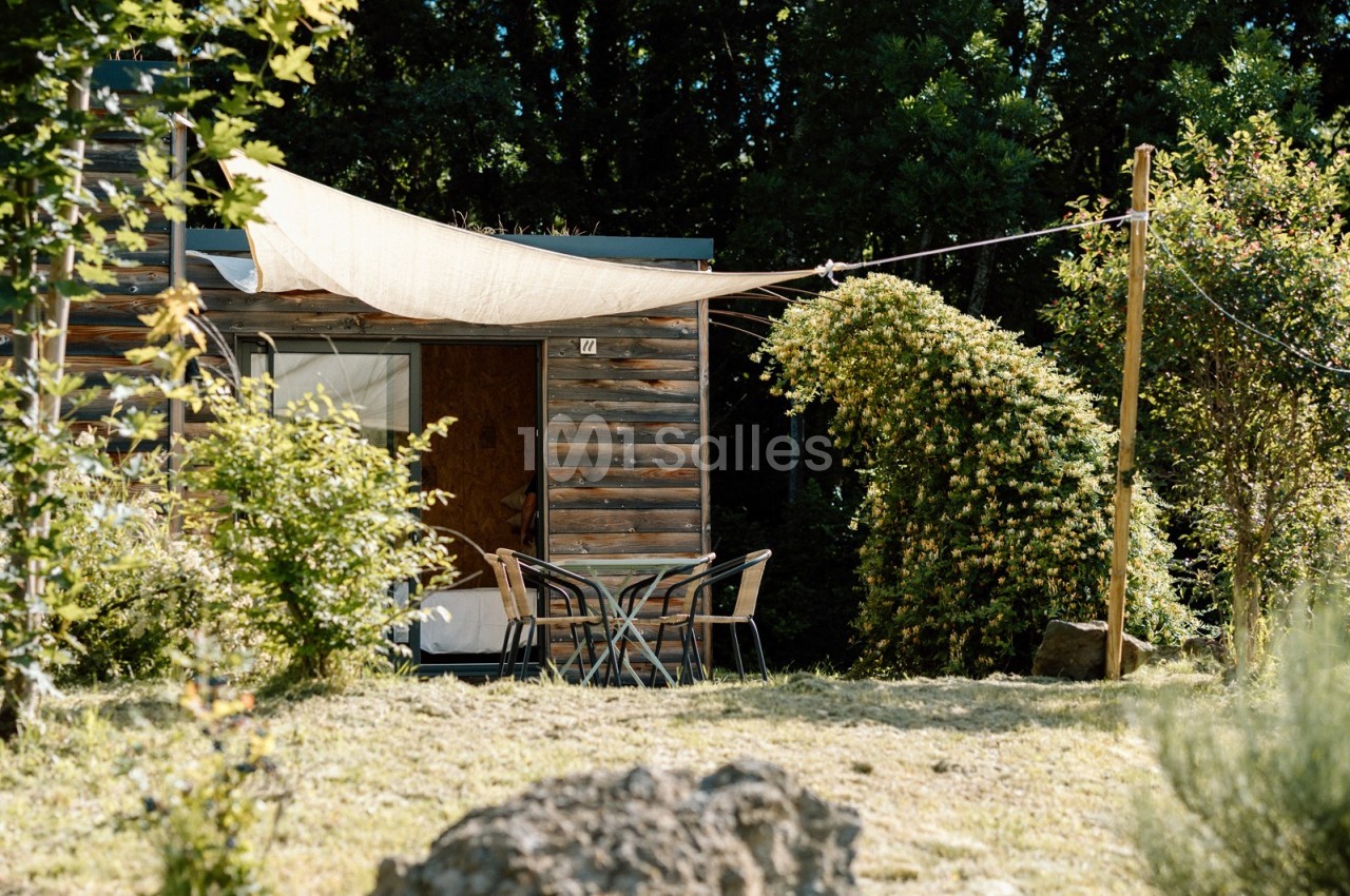 Table et chaises en extérieur devant une petite habitation en bois, entourée de végétation et ombragée par une toile tendue.