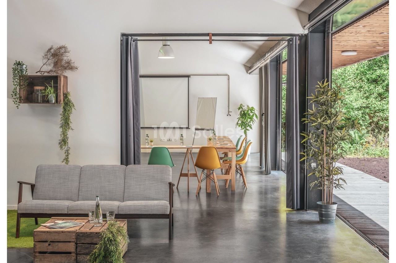 Salle lumineuse avec canapé, table en bois, chaises colorées, plantes, et grande ouverture sur un espace extérieur verdoyant.