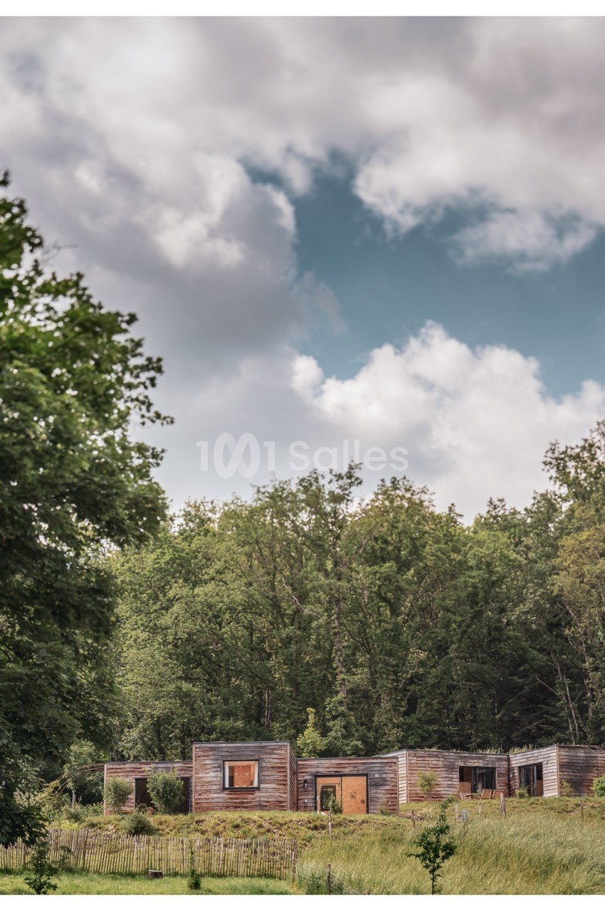 Maison en bois moderne entourée de verdure, avec un ciel partiellement nuageux en arrière-plan.