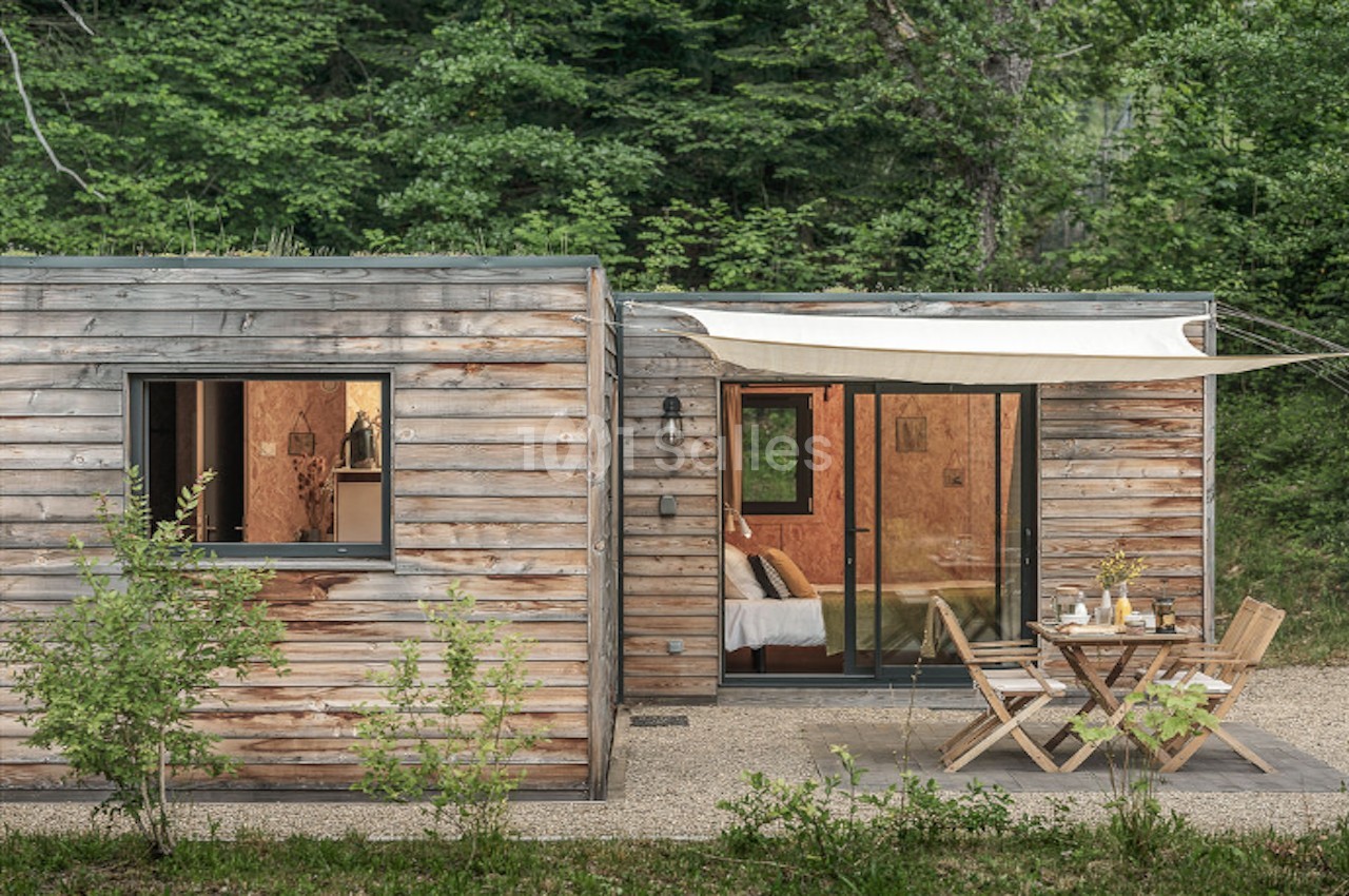 Cabane en bois avec terrasse aménagée, table et chaises, entourée de végétation dense en pleine nature.