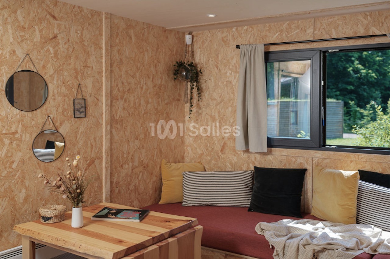 Salon lumineux avec murs en bois, canapé rouge, coussins colorés, table basse en bois et fenêtre donnant sur un jardin.