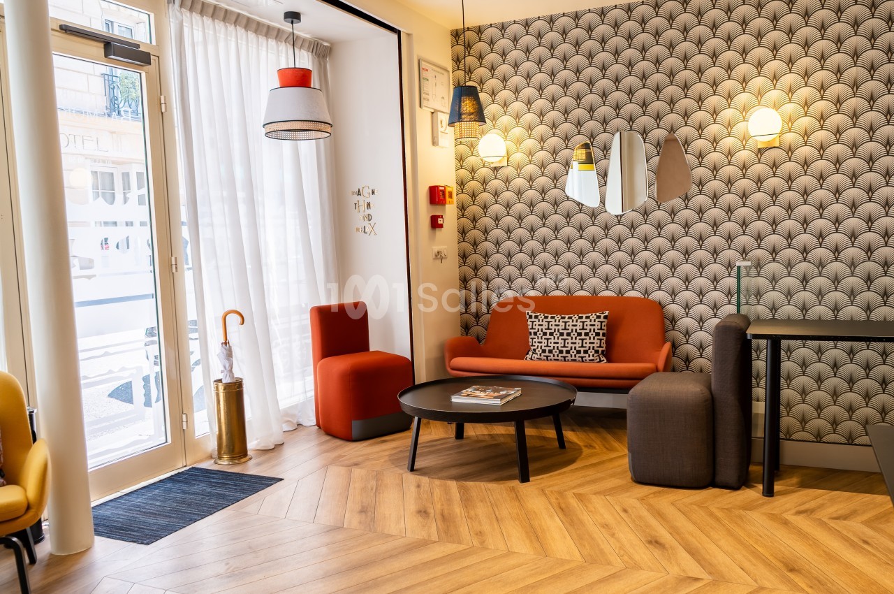Salon moderne avec canapé orange, table basse ronde, chaises, lampes suspendues et papier peint à motifs géométriques.