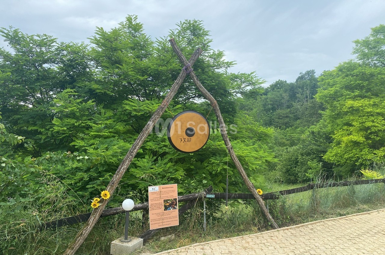 Un grand gong suspendu entre deux branches en triangle, entouré de verdure, près d'un chemin pavé.