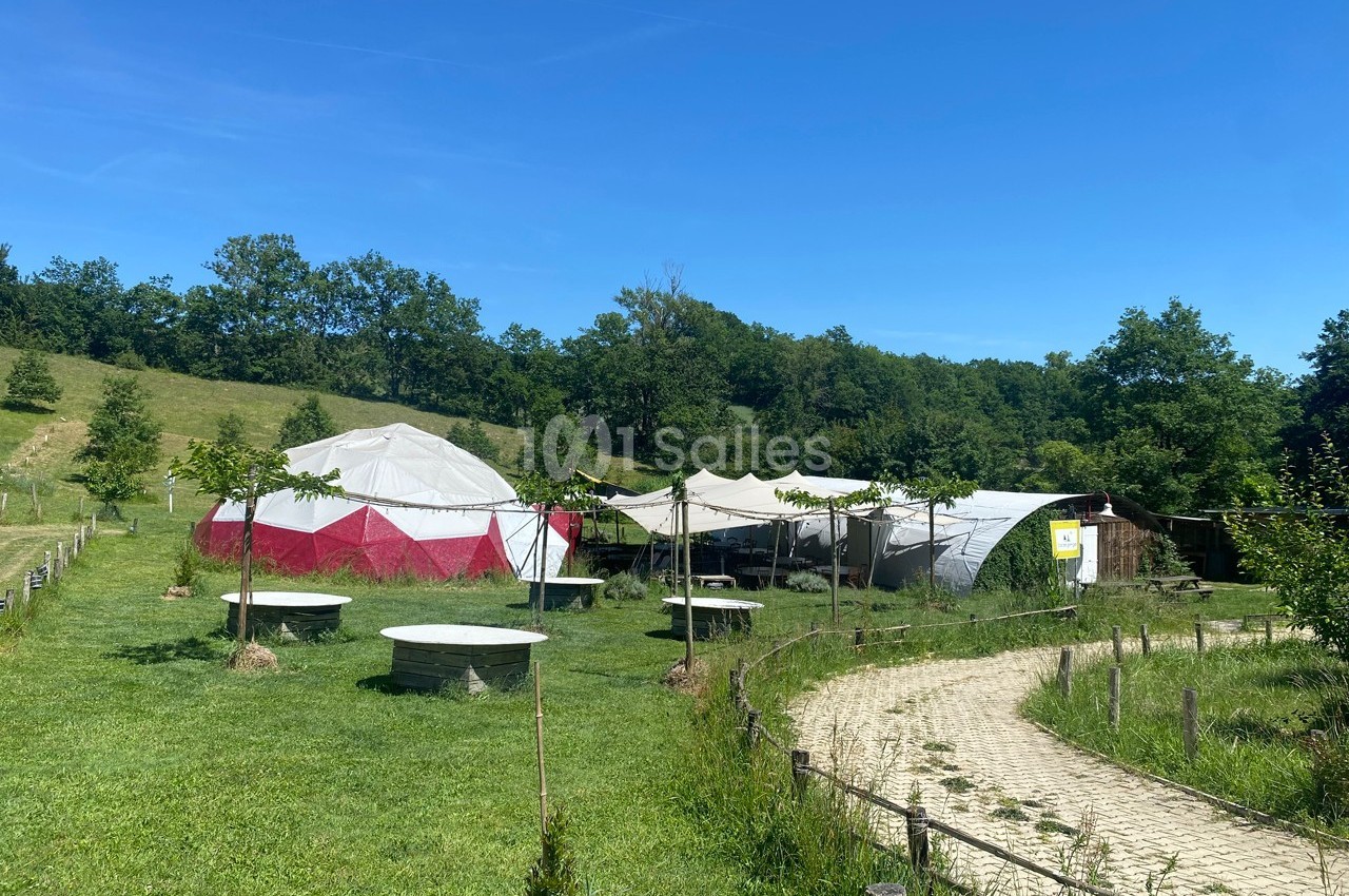 Deux grandes tentes et des tables en plein air dans un espace vert entouré d'arbres et de collines.