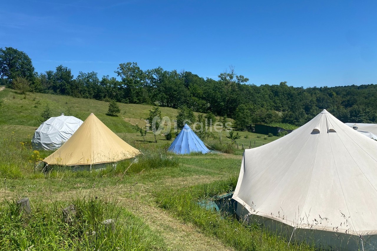 Tentes de camping installées sur une prairie verdoyante sous un ciel bleu, entourées d'arbres et de collines.