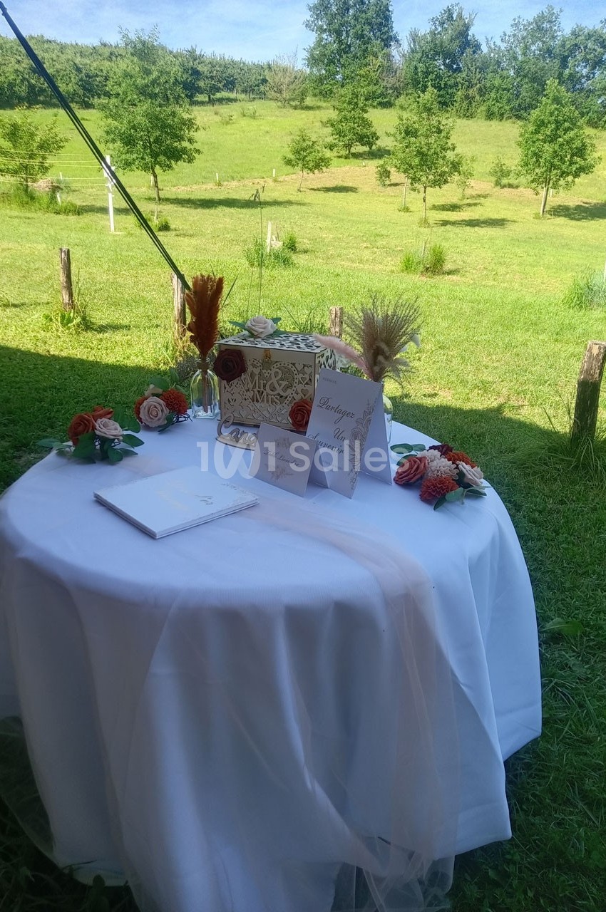 Table ronde décorée pour un mariage, avec urne, fleurs, cartes et vue sur un jardin verdoyant en arrière-plan.