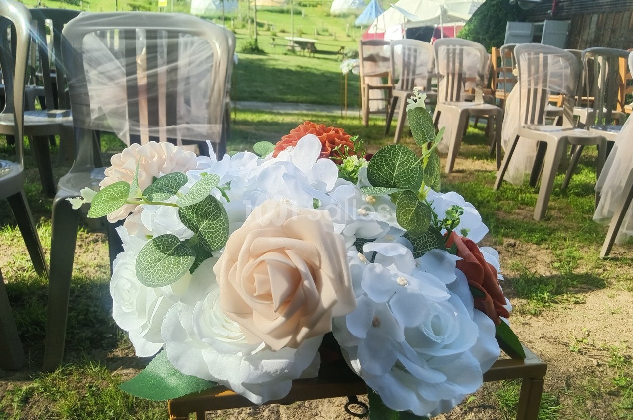Arrangement de fleurs blanches, beiges et orange sur une table, avec des chaises disposées en extérieur en arrière-plan.