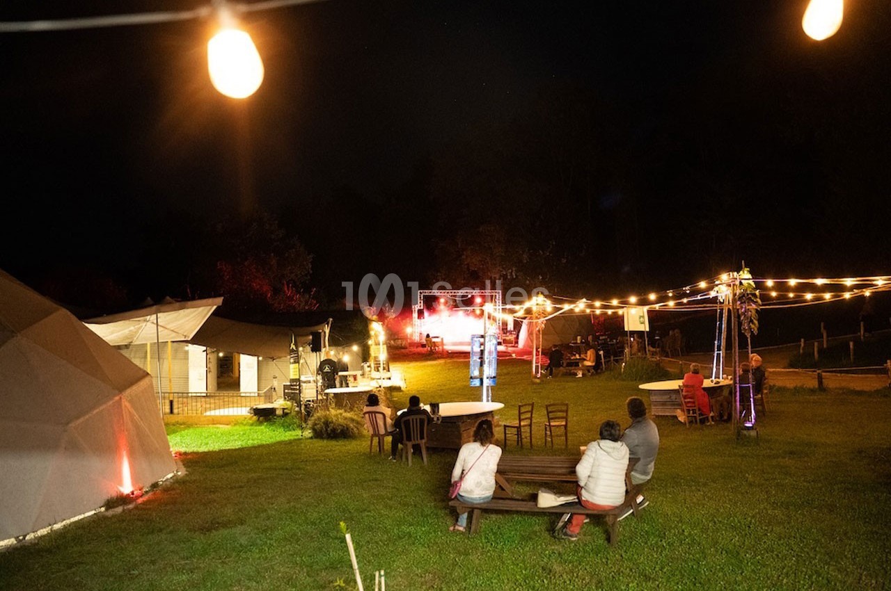 Espace extérieur éclairé de nuit avec des guirlandes lumineuses, des structures et des personnes assises sur des bancs.