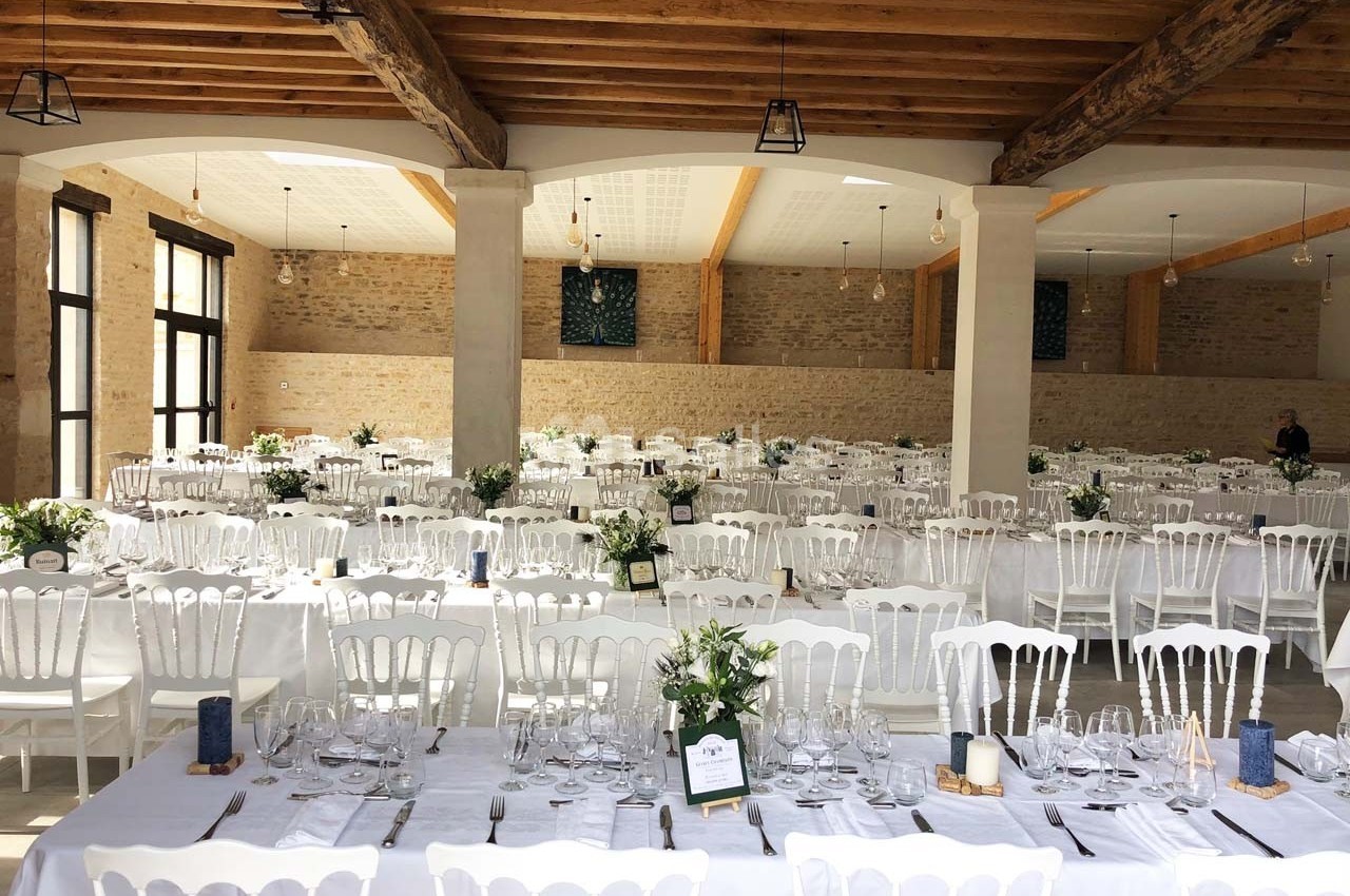 Salle de réception lumineuse avec de longues tables dressées, chaises blanches et décorations florales.