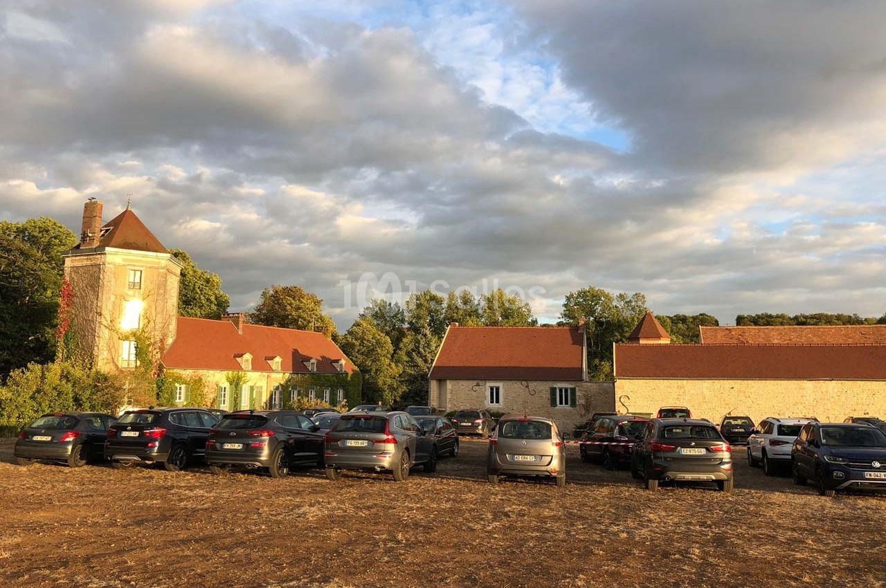 Parking de voitures devant des bâtiments anciens en pierre, sous un ciel partiellement nuageux en fin de journée.