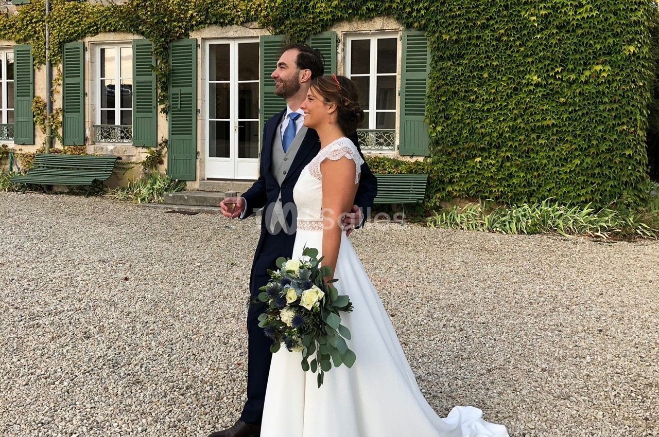 Un couple en tenue de mariage pose devant une maison en pierre couverte de lierre, sur une allée de gravier.