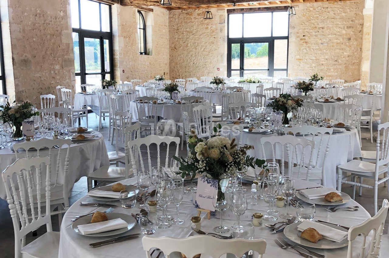 Salle de réception lumineuse avec tables rondes dressées, décorées de fleurs et couverts élégants.