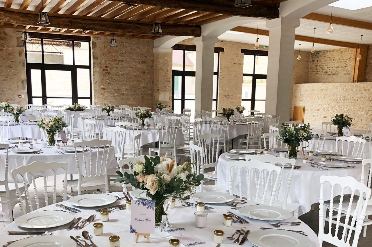 Salle de réception décorée avec des tables rondes, nappes blanches, chaises en bois blanc et bouquets floraux.