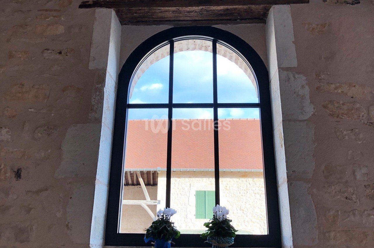 Fenêtre cintrée avec cadre noir, vue sur un toit en tuiles rouges et deux pots de fleurs blanches sur le rebord.