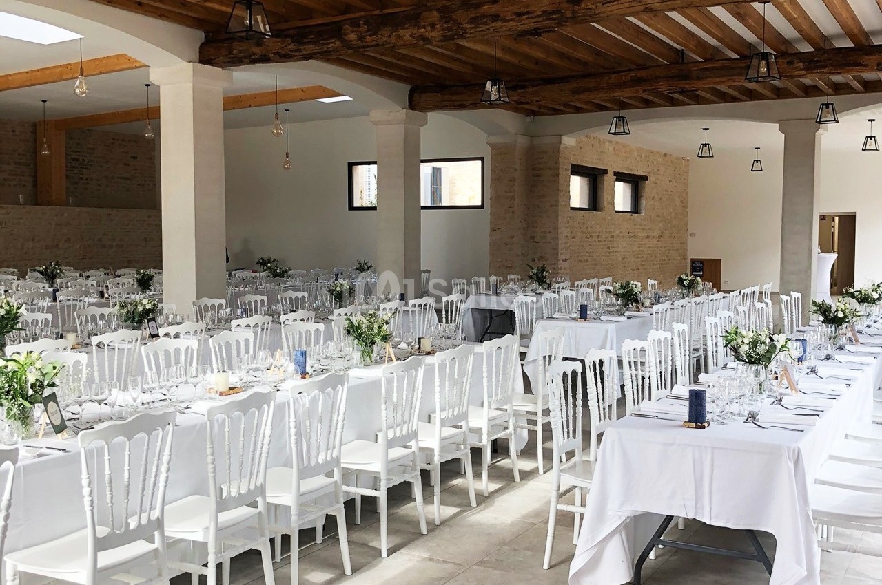 Salle de réception lumineuse avec de longues tables décorées de nappes blanches, chaises blanches et arrangements floraux.